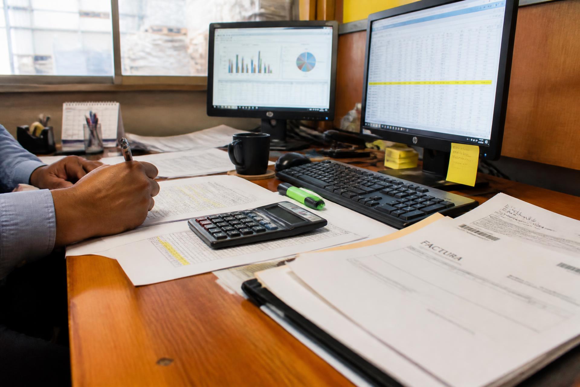 Escritorio de administración con planillas impresas, calculadora y pantallas con datos de ventas
